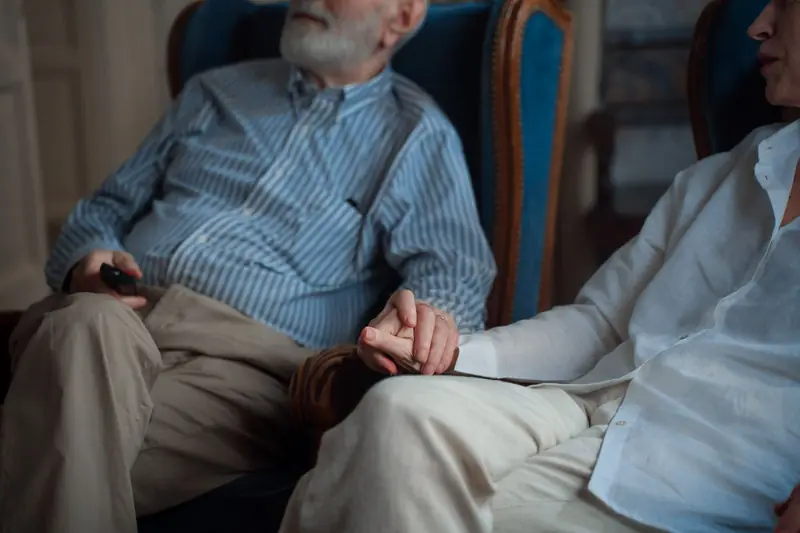 An older couple sit side by side in armchairs, holding hands