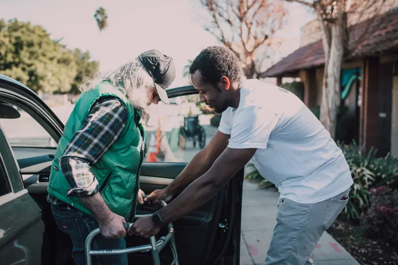 A young man helps an older man with a walker step out of a car